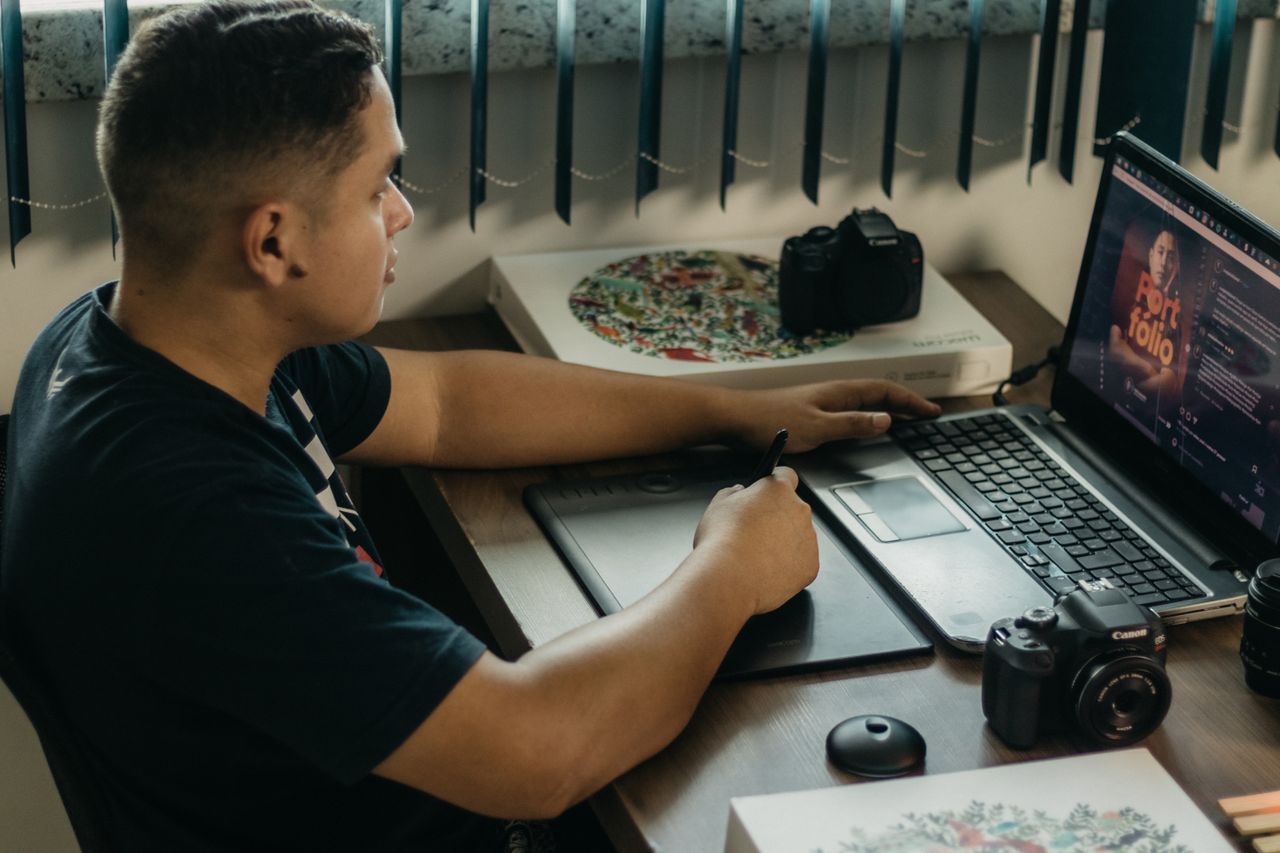 A man is editing photos on a laptop with a graphics tablet in an indoor workspace surrounded by camera gear.